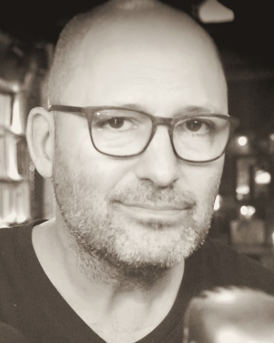 Portrait de Frédéric Bascuñana en noir et blanc chaud, lunettes, regard calme, fond de lumières floues.