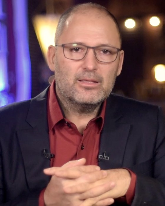 Frédéric Bascuñana en veste sombre et chemise rouge, mains jointes, sur un plateau de tournage aux lumières chaudes.